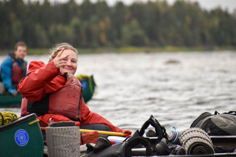 A woman in a red life jacket is sitting in a canoe, making a peace sign with her fingers. She is surrounded by gear, including bags and a sleeping mat. In the background, another person is visible in a canoe, and there is a line of trees along the shore. The water is choppy, and the sky is overcast.